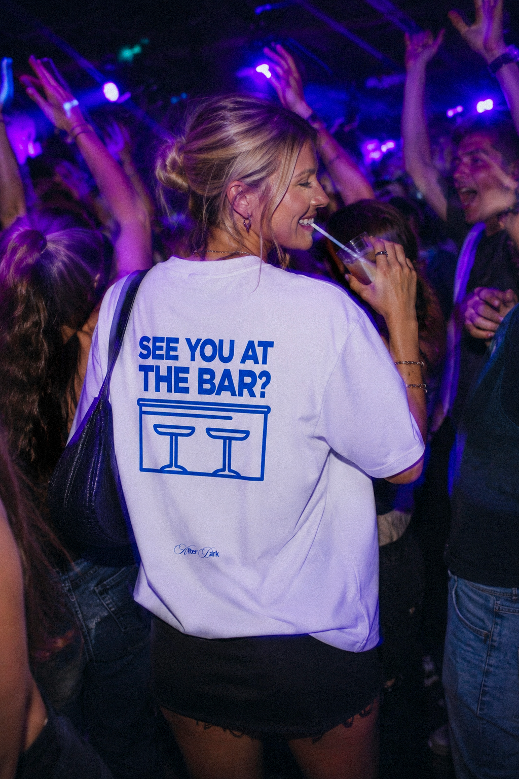 Modella indossa la t-shirt oversize bianca SEE YOU AT THE BAR? di 4AM Season in un club, con grafica blu sul retro