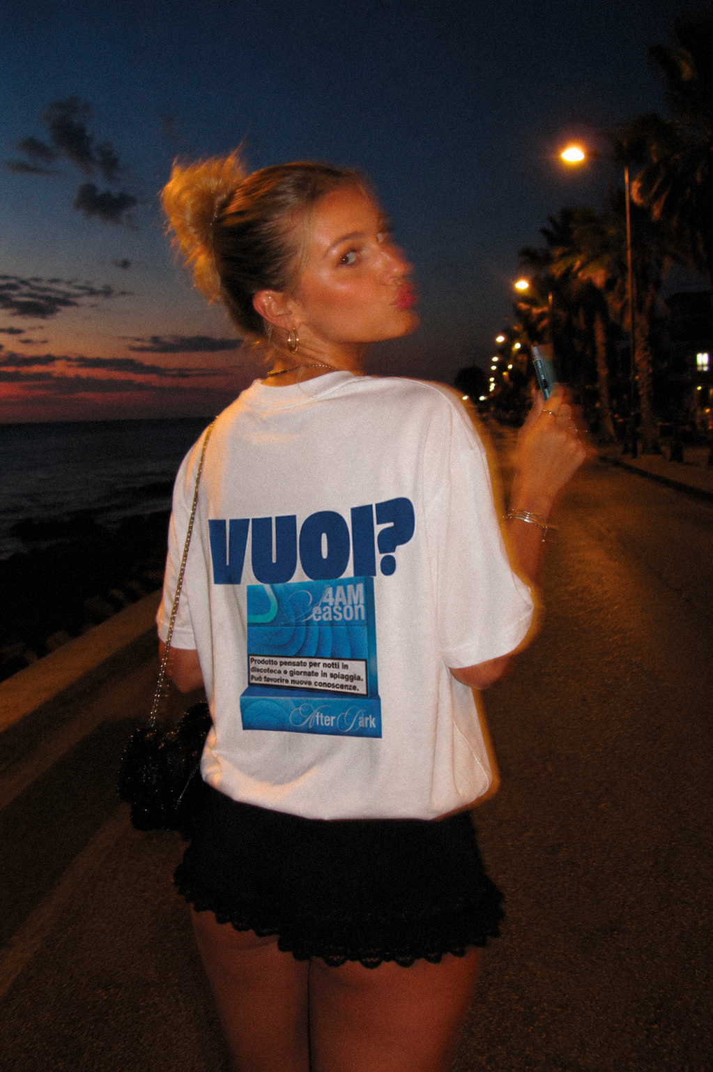 Modella indossa la t-shirt oversize bianca VUOI? di 4AM Season sul lungomare al tramonto, con grafica azzurra sul retro
